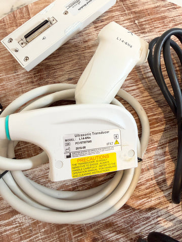 An ultrasound transducer probe with a coiled beige cable, a white plastic housing, and a black connector cable, placed on a wooden surface next to another white device.