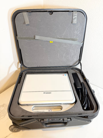 An open hard carrying case containing a white and silver Mindray portable ultrasound machine with a black accessory cord and strap inside a foam-lined compartment.