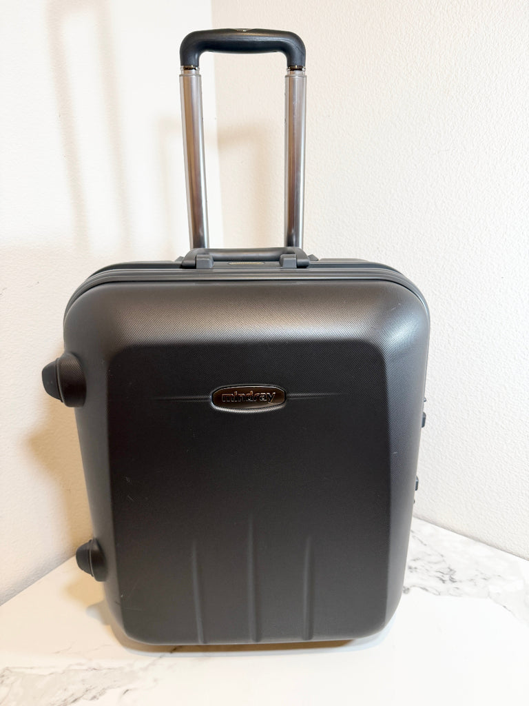 A black hard-shell carry-on suitcase with an extended telescoping handle, standing on a light marble-like surface against a white wall.