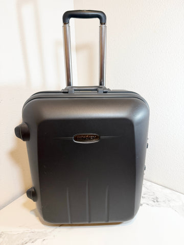 A black hard-shell carry-on suitcase with an extended telescoping handle, standing on a light marble-like surface against a white wall.