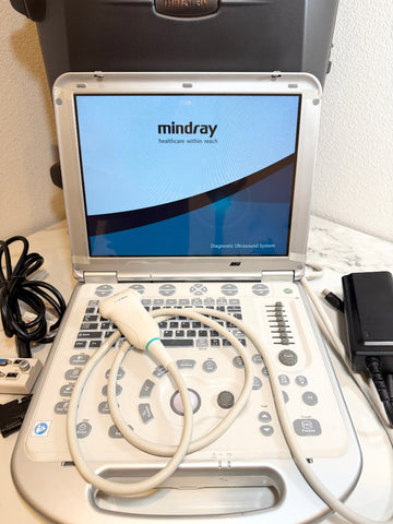 A Mindray portable ultrasound machine with its handpiece resting on the keyboard, set against a white textured wall with some cables and adapters visible.