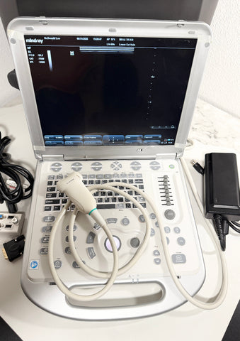A Mindray portable ultrasound machine with a white casing, featuring a screen, control panel, and attached ultrasound probe on a white surface, with cables and an external power adapter visible.