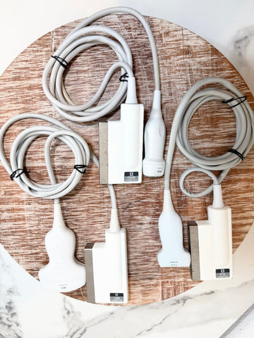 Three white and silver medical ultrasound transducers with coiled gray cables, arranged on a round wooden board over a white marble surface.
