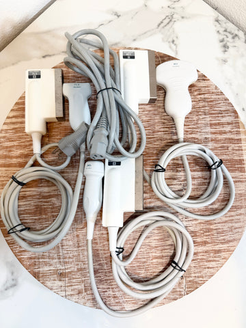 A set of medical ultrasound probes and cables laid out on a round wooden board, photographed from above on a white marble surface.