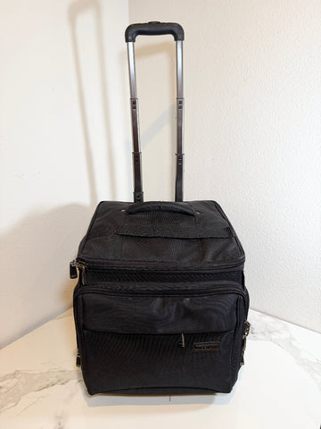 A black soft-sided rolling travel bag with an extended telescoping handle, standing upright on a light marble-like surface against a white wall.