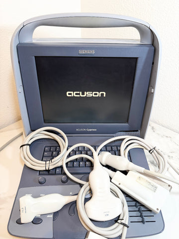 A Siemens Acuson ultrasound machine with its attached cables and probes displayed in front of the open monitor on a light surface against a textured wall.