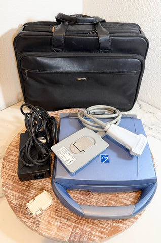A SonoSite MicroMaxx Portable Ultrasound System kit displayed on a round wooden tray, featuring a blue and gray ultrasound probe, a gray handpiece, a black power adapter with coiled cord, and a black carrying case in the background.