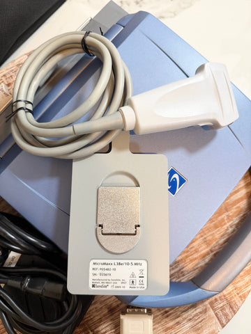 A medical ultrasound transducer probe with a coiled gray cable, resting on top of a blue and gray ultrasound machine. The probe has a white plastic handle and a gray mounting plate with a silver metal contact pad.