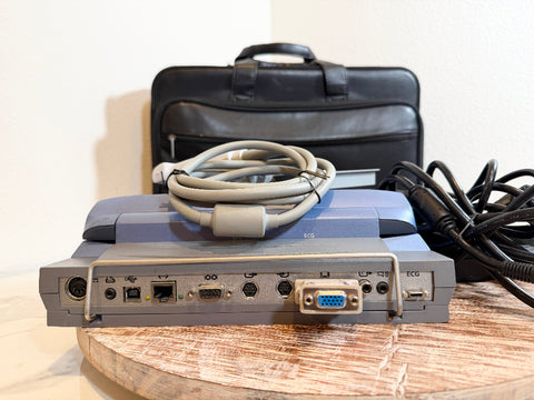 A SonoSite MicroMaxx Portable Ultrasound System with a gray front panel and blue top housing, shown with attached cables and a black carrying case on a wooden surface against a plain wall.