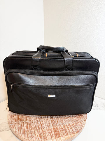 A black leather and nylon briefcase-style bag with top handles and a front zip pocket, displayed on a round wooden surface against a white wall corner.