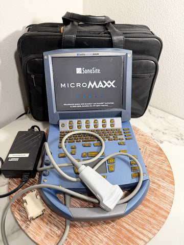 A SonoSite MicroMaxx handheld ultrasound machine with a blue and gray casing, attached cables, and a white probe, displayed on a round wooden tray with its black carrying case and power adapter behind it.