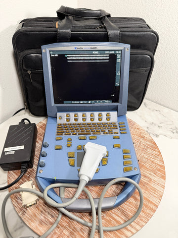 A blue SonoSite MicroMaxx Portable Ultrasound System with a keyboard, screen, and attached probe, shown with its black carrying case and power adapter on a round wooden tray against a white wall.