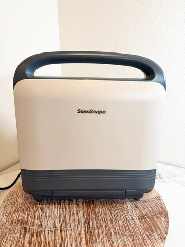 A SonoScape portable ultrasound machine with a cream-colored body, dark gray handle and base, sitting on a wooden surface against a white wall.