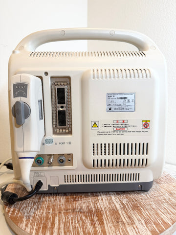 A portable Sonoscape ultrasound machine, shown from the back with its handle, connectors, and power cord visible, sitting on a wooden surface against a plain wall.