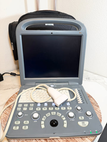 A Sonoscape ultrasound machine with a gray console, black screen, and attached white probe cable, placed on a round wooden tray with a black carrying case behind it.