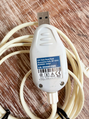 A Welch Allyn SpiroPerfect 703554 USB Spirometer Sensor white USB cable adapter with a coiled cream-colored cable, photographed on a brown wood-grain surface.