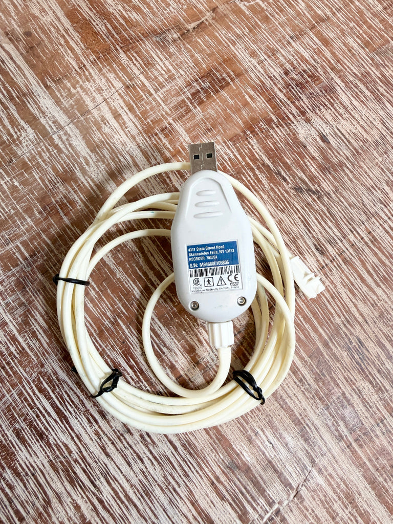 A white USB adapter cable with a built-in toggle switch, coiled on a brown wood-grain surface. The product features a white plastic body with a blue and white label, a USB connector on one end, and a white plug on the other.