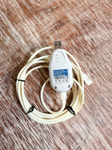 A white USB adapter cable with a built-in toggle switch, coiled on a brown wood-grain surface. The product features a white plastic body with a blue and white label, a USB connector on one end, and a white plug on the other.