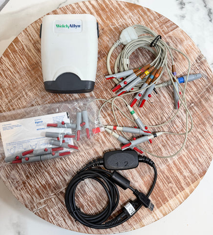 Welch Allyn ECG/ECG-EEG electrode kit with multiple gray and red electrode leads, a white and gray device, a black cable with a USB connector, and a plastic bag of electrode connectors, all arranged on a round wooden board.