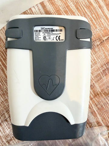 A handheld EKG/ECG monitor device photographed from the back, featuring a white body with dark gray top and bottom sections, a central heart-shaped emblem with an ECG line, and a product label on the upper back. The device is placed on a textured brown surface.