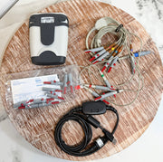 A medical-grade ECG/ECG monitor kit laid out on a round wooden board, including a white and black monitor unit, multiple gray and red electrode leads, a black cable with a USB connector, a plastic bag of electrode connectors, and printed paperwork.