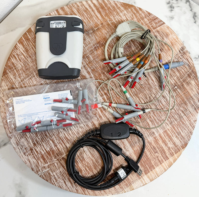A medical-grade ECG/ECG monitor kit laid out on a round wooden board, including a white and black monitor unit, multiple gray and red electrode leads, a black cable with a USB connector, a plastic bag of electrode connectors, and printed paperwork.