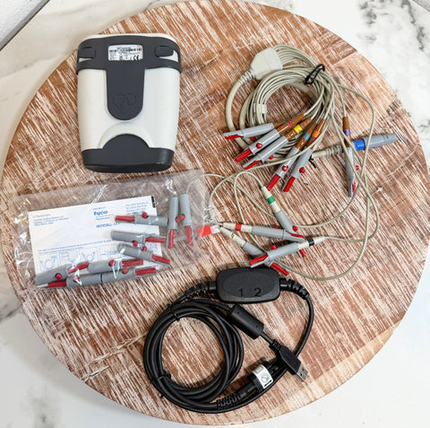 A medical-grade ECG/ECG monitor kit laid out on a round wooden board, including a white and black monitor unit, multiple gray and red electrode leads, a black cable with a USB connector, a plastic bag of electrode connectors, and printed paperwork.