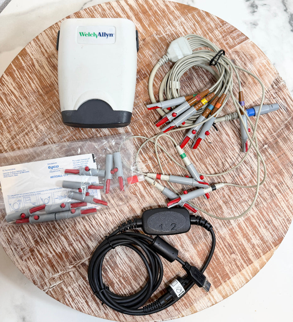 Welch Allyn ECG/ECG electrode kit laid out on a round wooden board, including a white and gray power unit, multiple gray and red electrode leads, a black cable with connectors, and a plastic bag of replacement electrode tips.